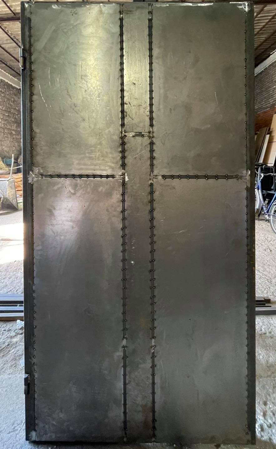 Duże, metalowe drzwi warsztatowe, spawane z blach, widoczne ślady spawania i nity, w tle warsztat z narzędziami i rowerem.