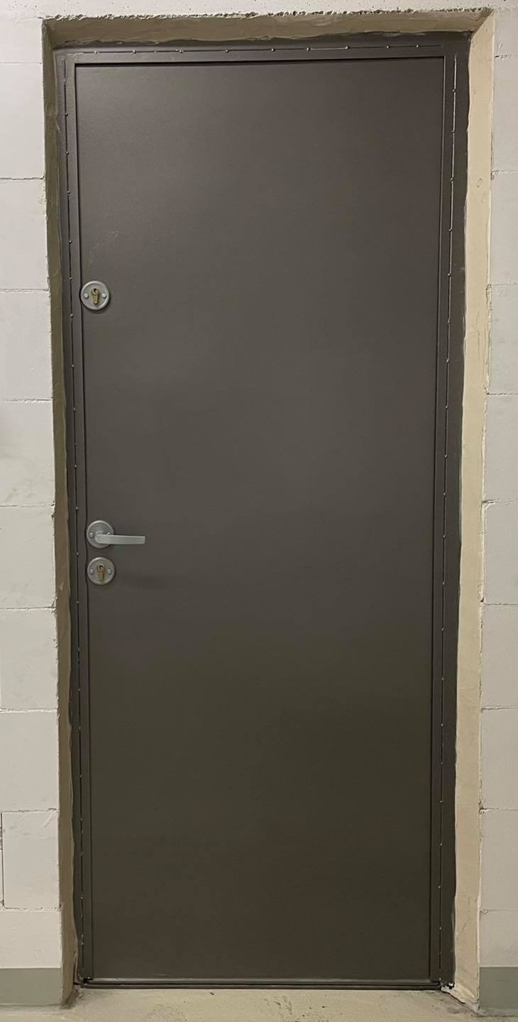Szare, stalowe drzwi wejściowe z zamkiem i klamką, osadzone w surowej, białej ścianie z betonu komórkowego.