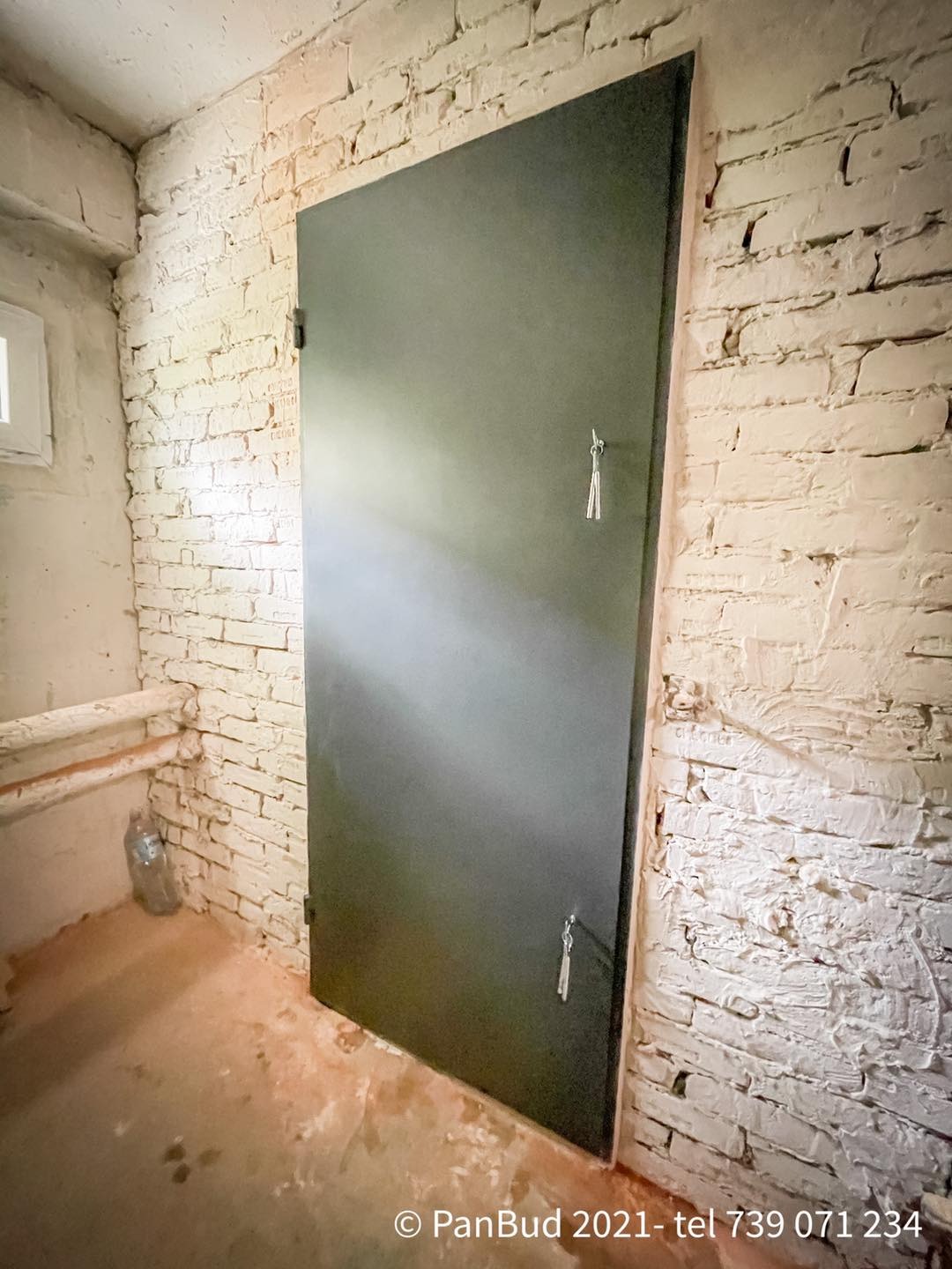 Ciemna, metalowa brama w piwnicy z surowymi, pomalowanymi na biało ceglanymi ścianami i betonową podłogą w trakcie remontu.