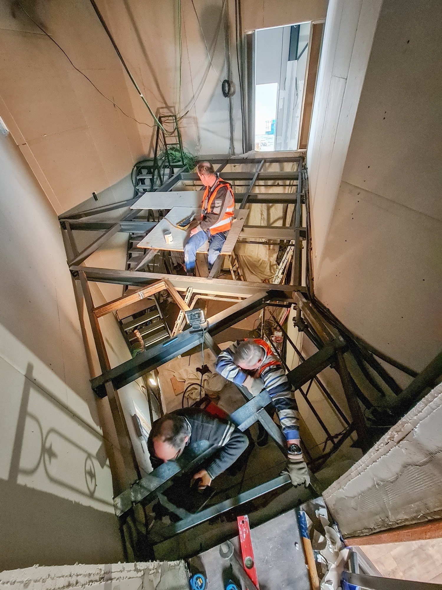 Trzech pracowników w kamizelkach odblaskowych montuje metalową konstrukcję pod sufitem w pomieszczeniu o surowych ścianach. Jeden siedzi na konstrukcji, drugi pracuje na niższym poziomie, a trzeci...