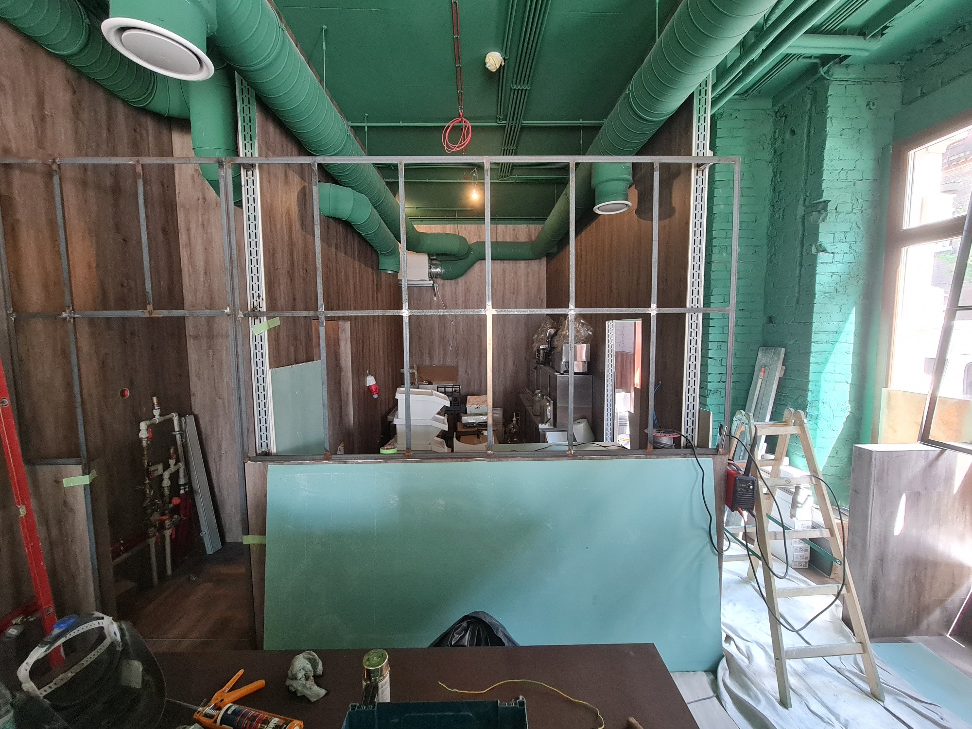 Wnętrze w trakcie remontu: widoczne metalowe profile ścianki działowej, zielone rury wentylacyjne pod sufitem, surowe ściany z cegły i drewnianych paneli, drabina i narzędzia malarskie.