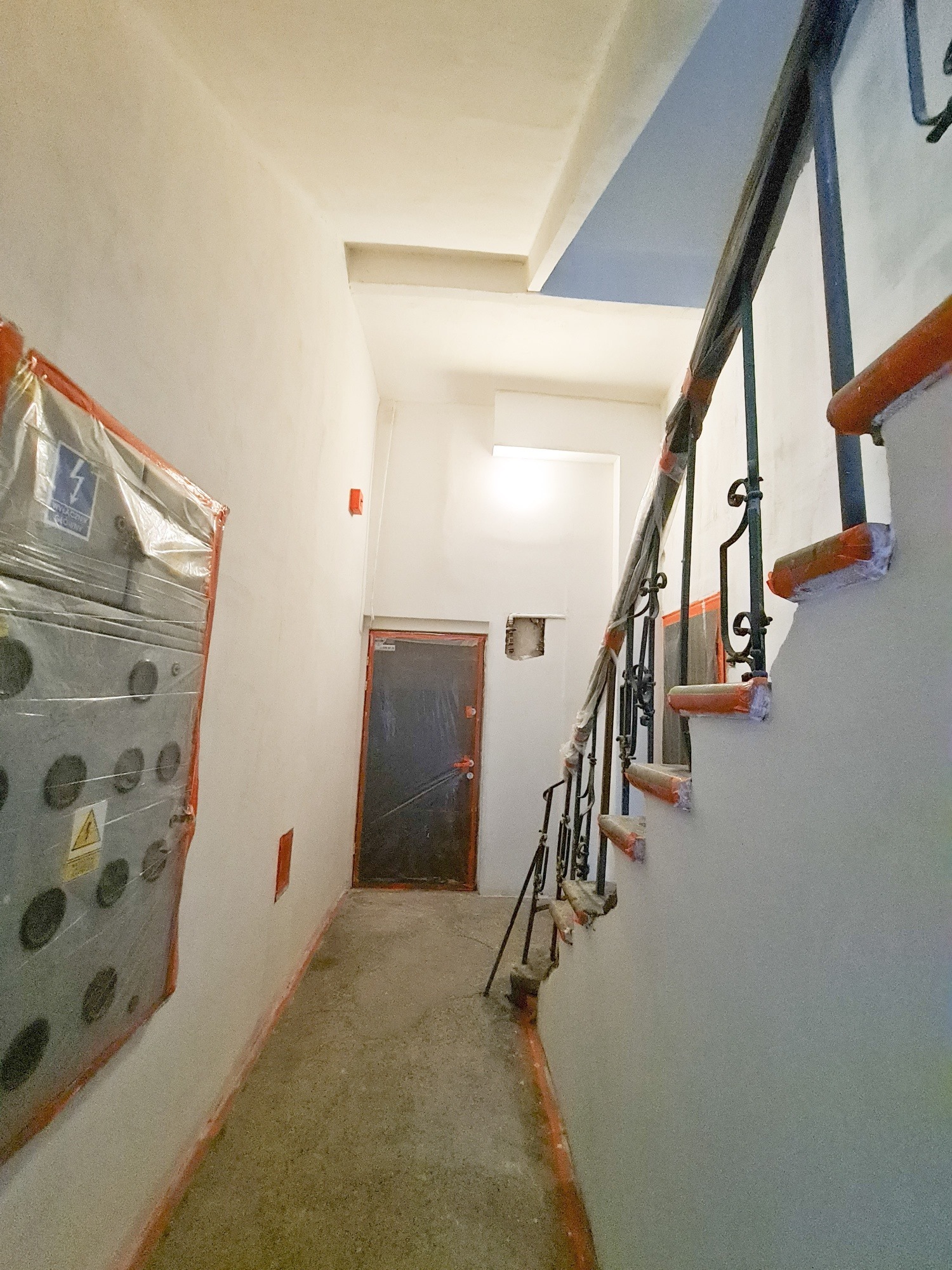 Korytarz w trakcie remontu: ściany przygotowane do malowania, zabezpieczona folią szafka elektryczna, schody z zabezpieczonymi stopniami, widoczne ślady po pracach.