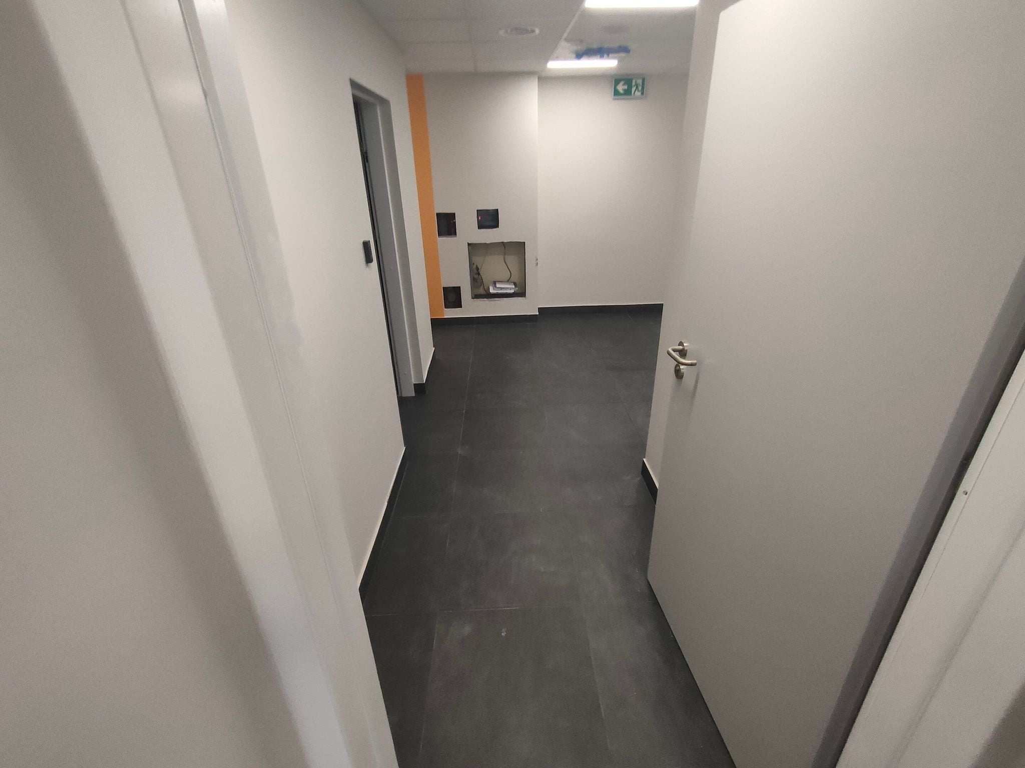 Korytarz biurowy z czarną podłogą, białymi ścianami i otwartymi drzwiami, widoczna wnęka z instalacją elektryczną i pomarańczowy akcent na ścianie.