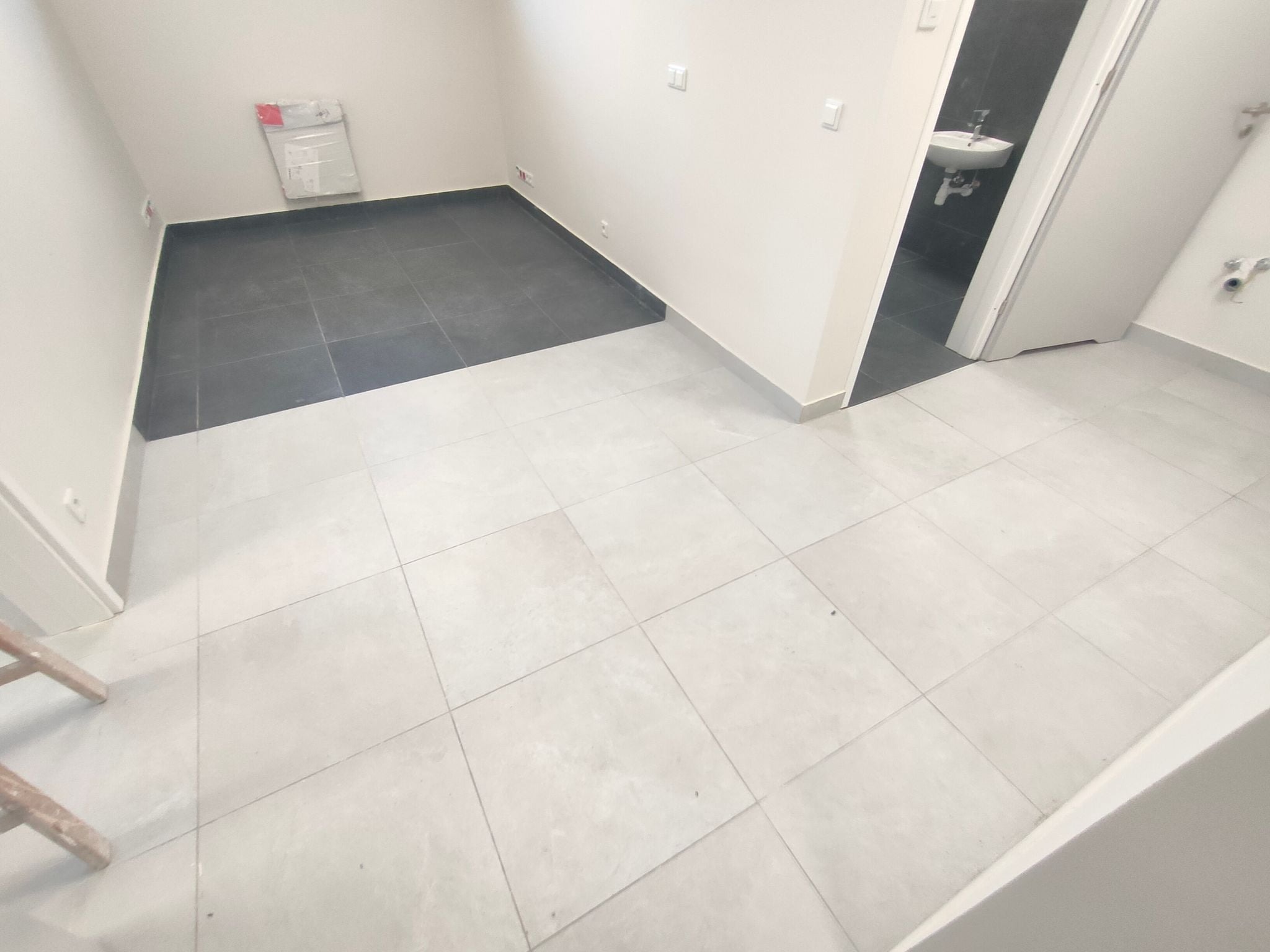 Wykończone wnętrze z dwoma rodzajami płytek podłogowych: ciemnoszarymi w części pomieszczenia i jasnoszarymi w pozostałej części, z widocznym wejściem do łazienki z umywalką.