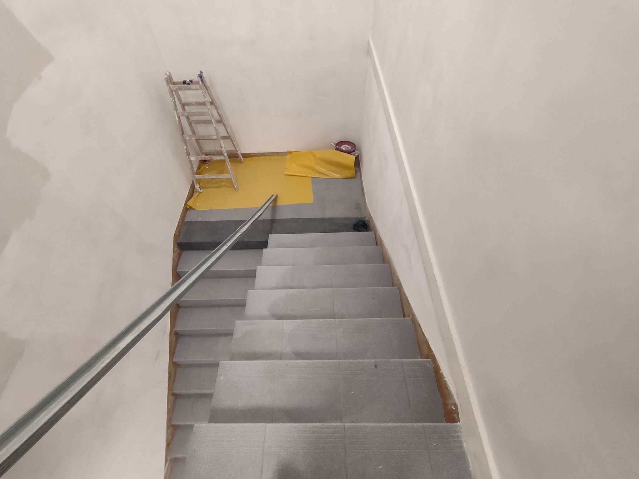 Klatka schodowa w trakcie remontu, zabezpieczona folią malarską, widoczna drabina oparta o ścianę i listwa przypodłogowa.
