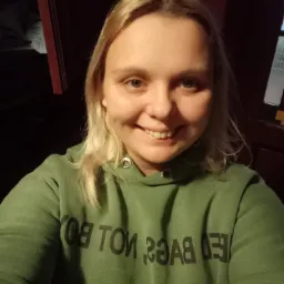 Uśmiechnięta blondynka w zielonej bluzie z kapturem i napisem 'NOT BOY' wykonuje selfie w pomieszczeniu z ciemnymi meblami.