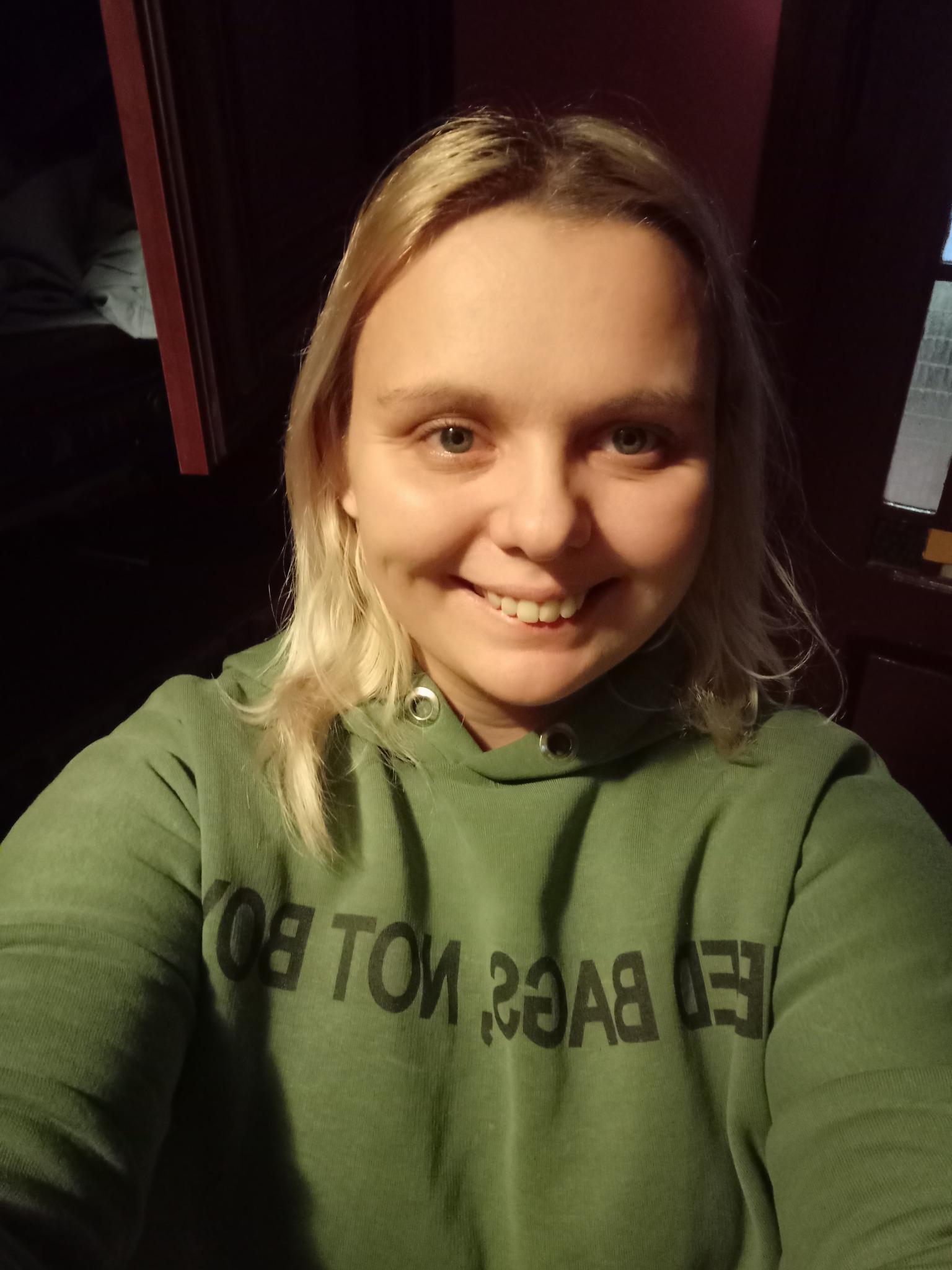 Uśmiechnięta blondynka w zielonej bluzie z kapturem i napisem 'NOT BOY' wykonuje selfie w pomieszczeniu z ciemnymi meblami.