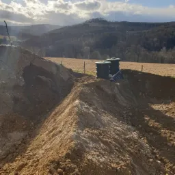 Dwa zbiorniki z zielonymi pokrywami połączone rurami, umieszczone w wykopie ziemnym na tle pagórkowatego krajobrazu pod pochmurnym niebem.