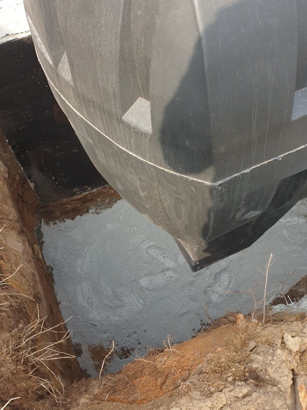 Częściowo zanurzony, czarny, plastikowy zbiornik w wykopie wypełnionym szarą, płynną substancją, prawdopodobnie betonem lub zaprawą, widoczne ślady po mieszaniu, ziemia na krawędzi wykopu.