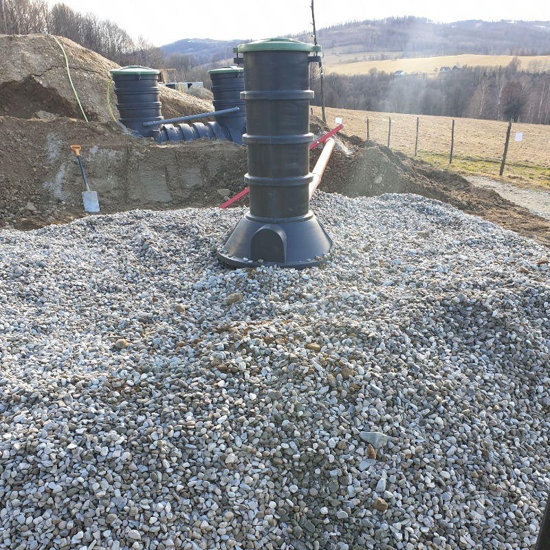 Montaż instalacji: czarny zbiornik z rurami na warstwie jasnych kamieni, w tle ziemny wykop z widocznymi elementami instalacji i krajobraz wzgórz.