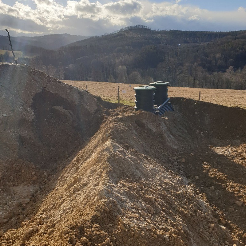 Dwa zbiorniki z zielonymi pokrywami połączone rurami, umieszczone w wykopie ziemnym na tle pagórkowatego krajobrazu pod pochmurnym niebem.