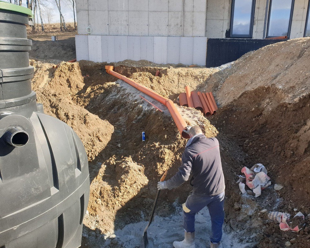 Montaż instalacji kanalizacyjnej: pracownik w wykopie z łopatą, widoczne rury kanalizacyjne pomarańczowe, zbiornik retencyjny z tworzywa sztucznego, fragment fundamentu budynku w tle.