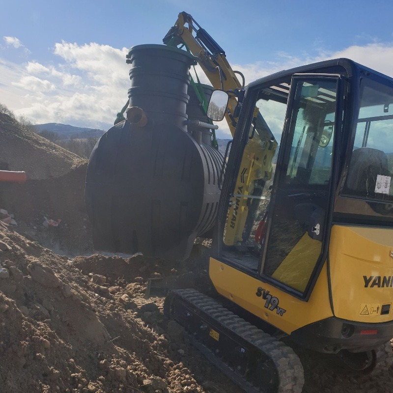 Żółta mini koparka Yanmar transportuje czarny, dwukomorowy zbiornik z tworzywa sztucznego na tle wykopu i błękitnego nieba z chmurami.