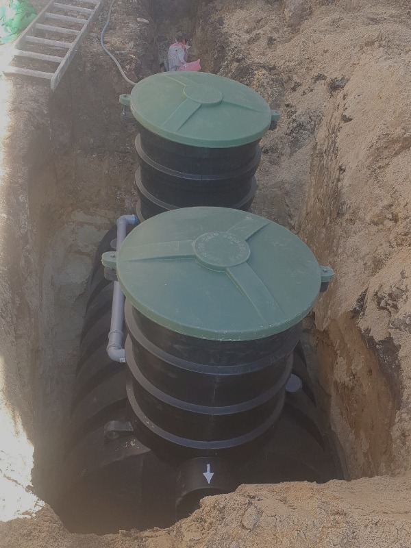 Dwu-komorowy, czarny zbiornik z zielonymi pokrywami, umieszczony w wykopie z piaszczystą ziemią, widoczna drabina oparta o krawędź wykopu.