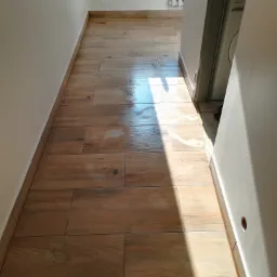 Korytarz z nową podłogą wyłożoną brązowymi płytkami imitującymi drewno, z widocznym fragmentem pralki w tle i silnym światłem słonecznym padającym na podłogę.