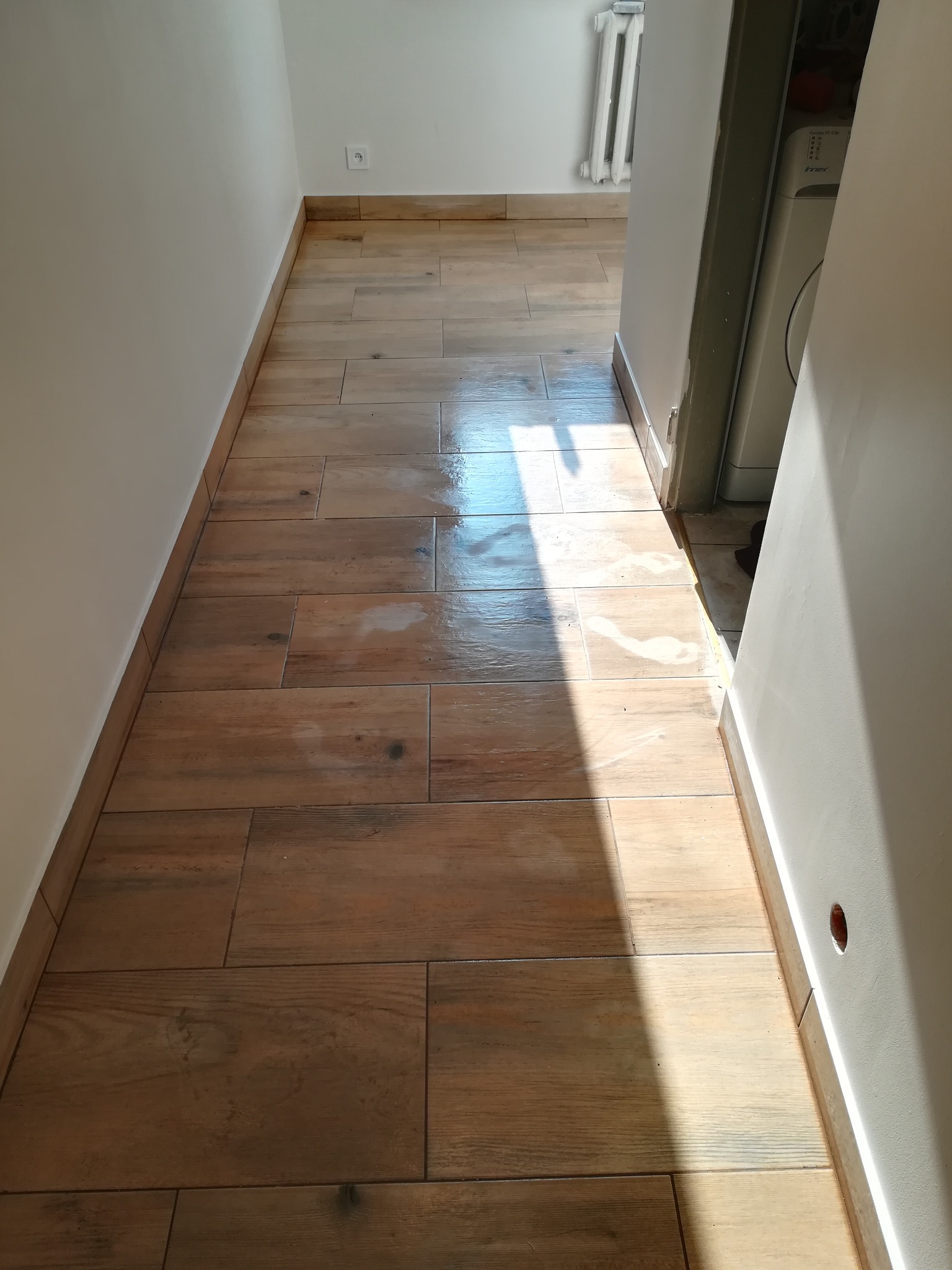 Korytarz z nową podłogą wyłożoną brązowymi płytkami imitującymi drewno, z widocznym fragmentem pralki w tle i silnym światłem słonecznym padającym na podłogę.