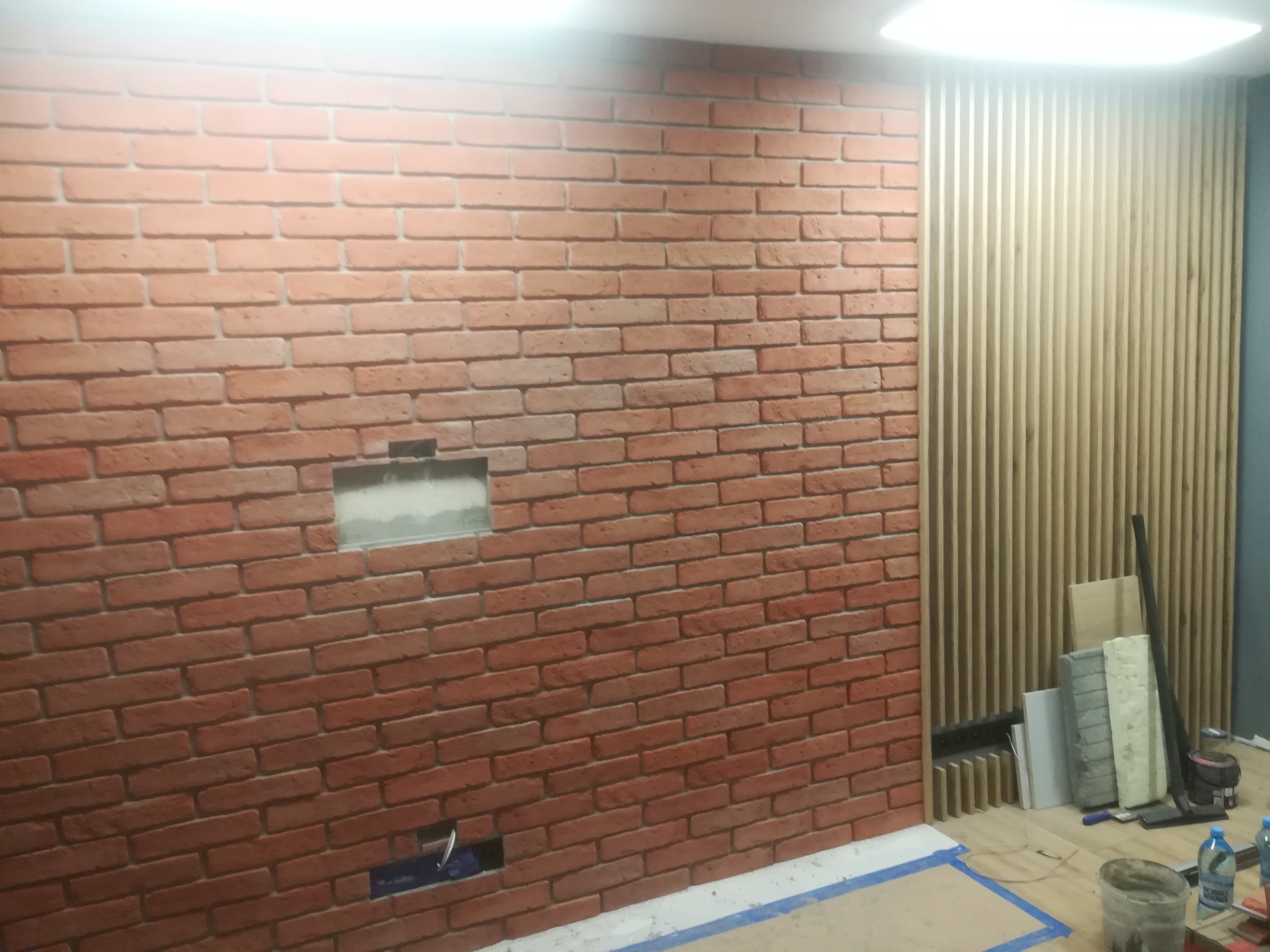 Ściana z czerwonej cegły z przygotowanymi otworami na instalacje, sąsiadująca z panelami z drewnianych listewek, widoczne materiały i narzędzia budowlane na podłodze.