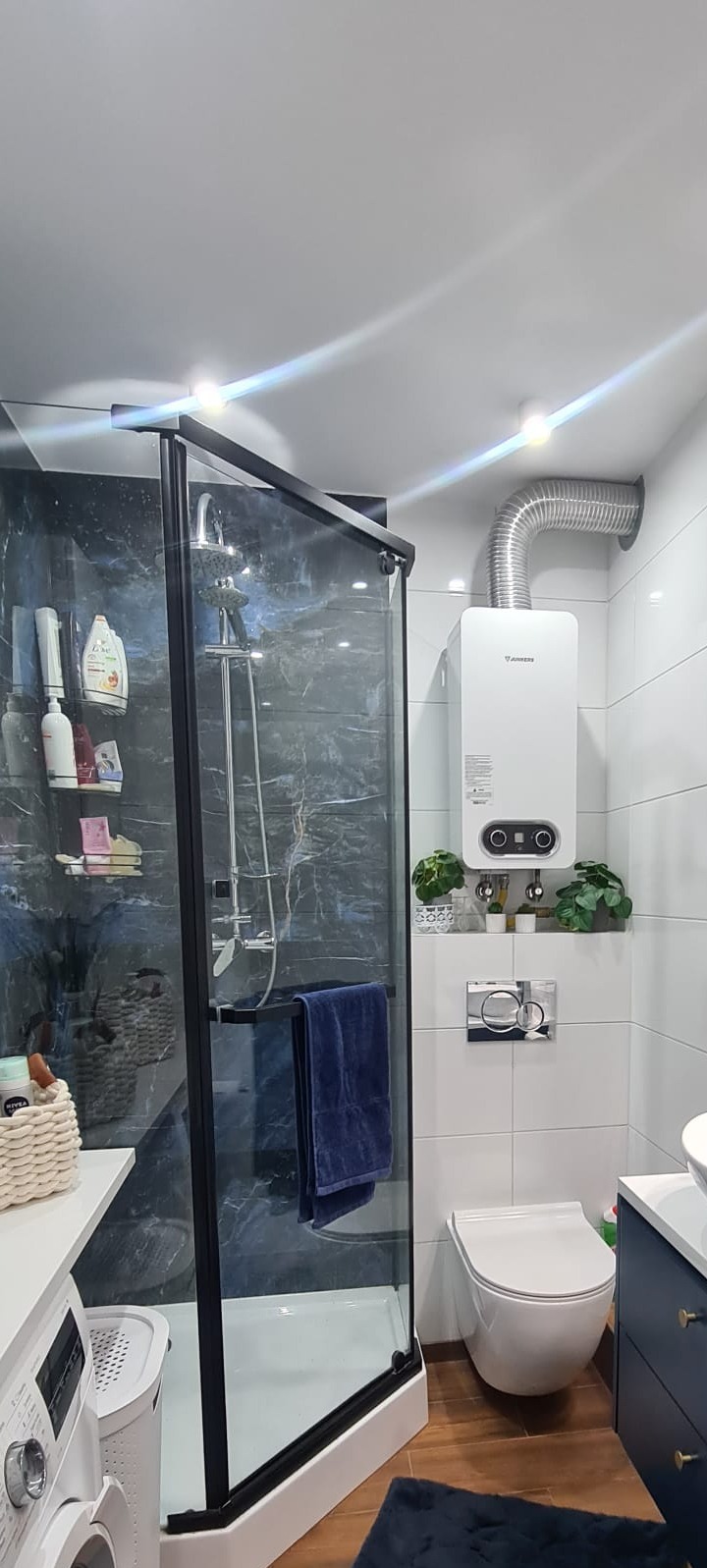 Nowoczesna łazienka z kabiną prysznicową w czarnej ramie, marmurowymi płytkami na ścianie prysznica, wiszącą toaletą, grzejnikiem gazowym Junkers i drewnopodobną podłogą.
