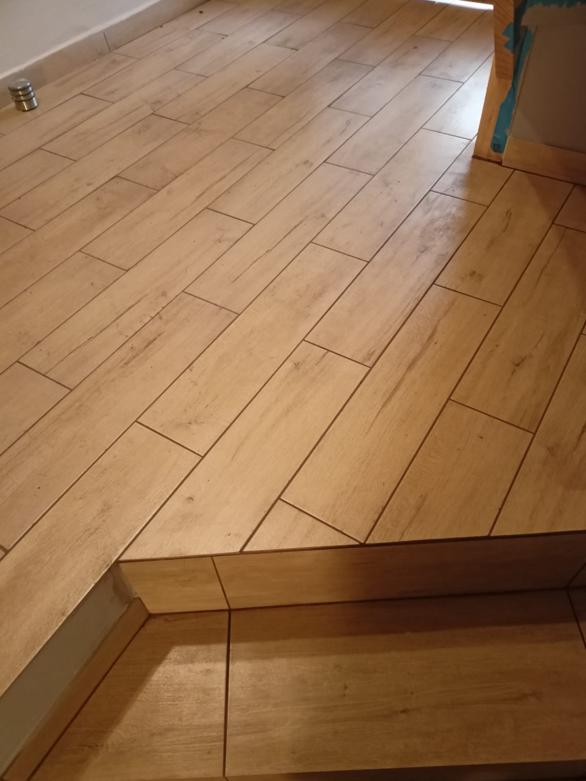 Beżowe, drewnopodobne płytki gresowe ułożone na podłodze i schodku, z widoczną fugą i listwą przypodłogową.