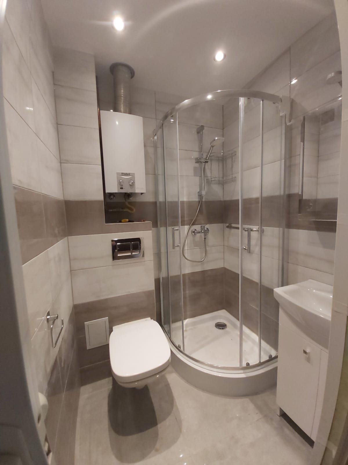 Nowoczesna łazienka po renowacji z kabiną prysznicową z półokrągłymi drzwiami, podwieszaną toaletą, umywalką z szafką i piecem gazowym na ścianie wyłożonej płytkami w odcieniach bieli i beżu.