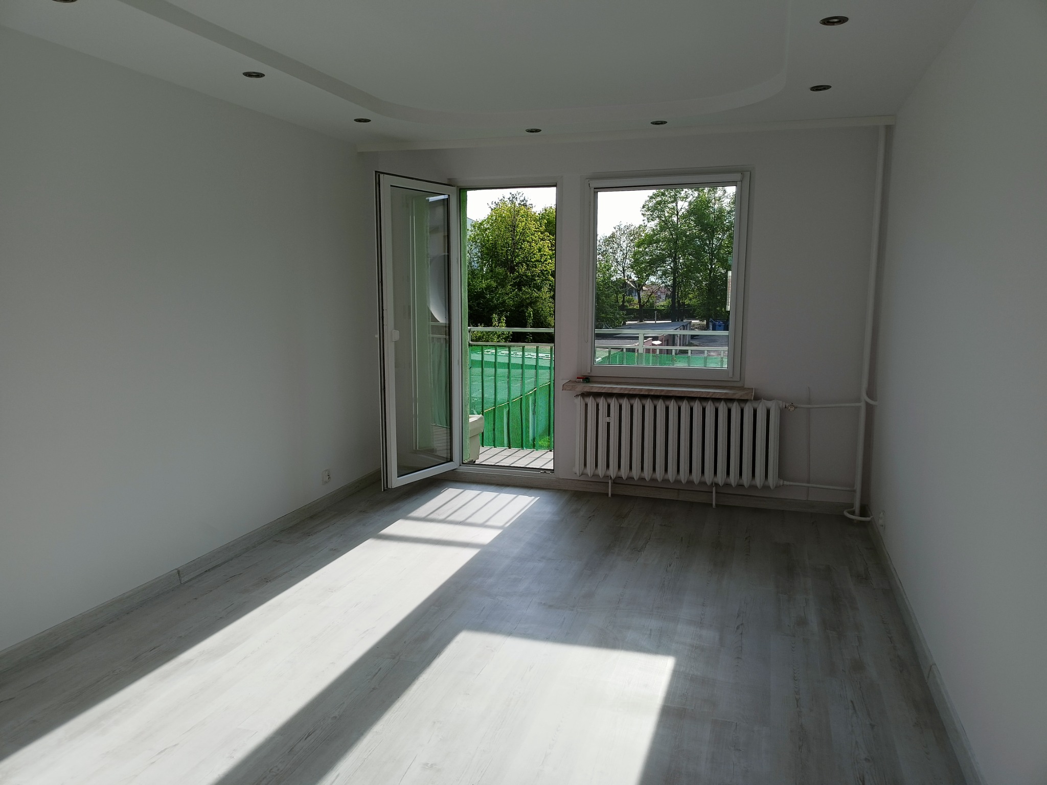 Pusty, jasny pokój po remoncie z panelami podłogowymi imitującymi drewno, białymi ścianami, otwartym balkonem z zieloną barierką i widokiem na drzewa, oraz grzejnikiem pod oknem.