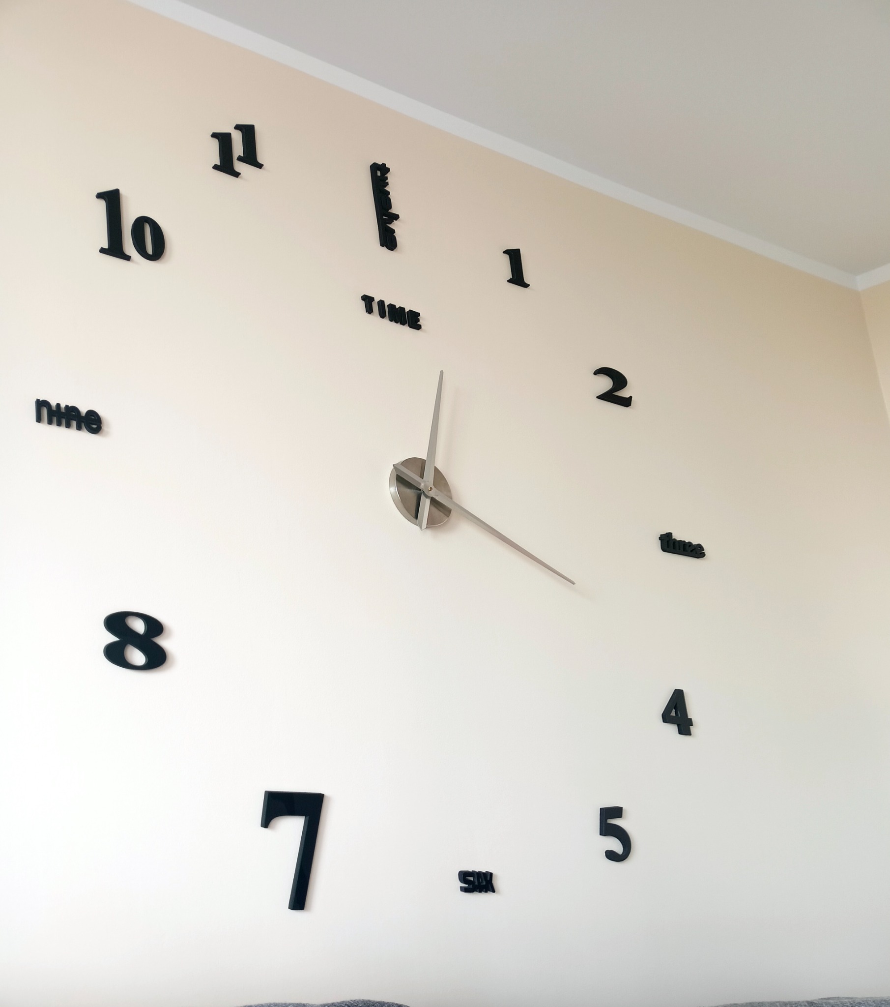 Duży, samoprzylepny zegar ścienny z czarnymi cyframi i srebrnymi wskazówkami na jasnej ścianie.
