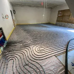 Rozłożone rury ogrzewania podłogowego na folii z siatką, widoczne kolektory z rurami w różnych kolorach oraz niedokończone ściany i sufit pomieszczenia.