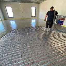 Wylewka anhydrytowa w trakcie realizacji: pracownicy rozprowadzają płynny jastrych na ogrzewanie podłogowe, widoczne okna w tle i metalowa konstrukcja sufitu.