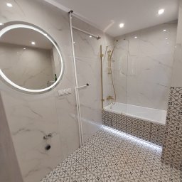 Szymon Zaworski - Nowoczesna łazienka z marmurowymi płytkami, okrągłym lustrem z podświetleniem LED, złotą armaturą prysznicową i podświetlaną wanną.