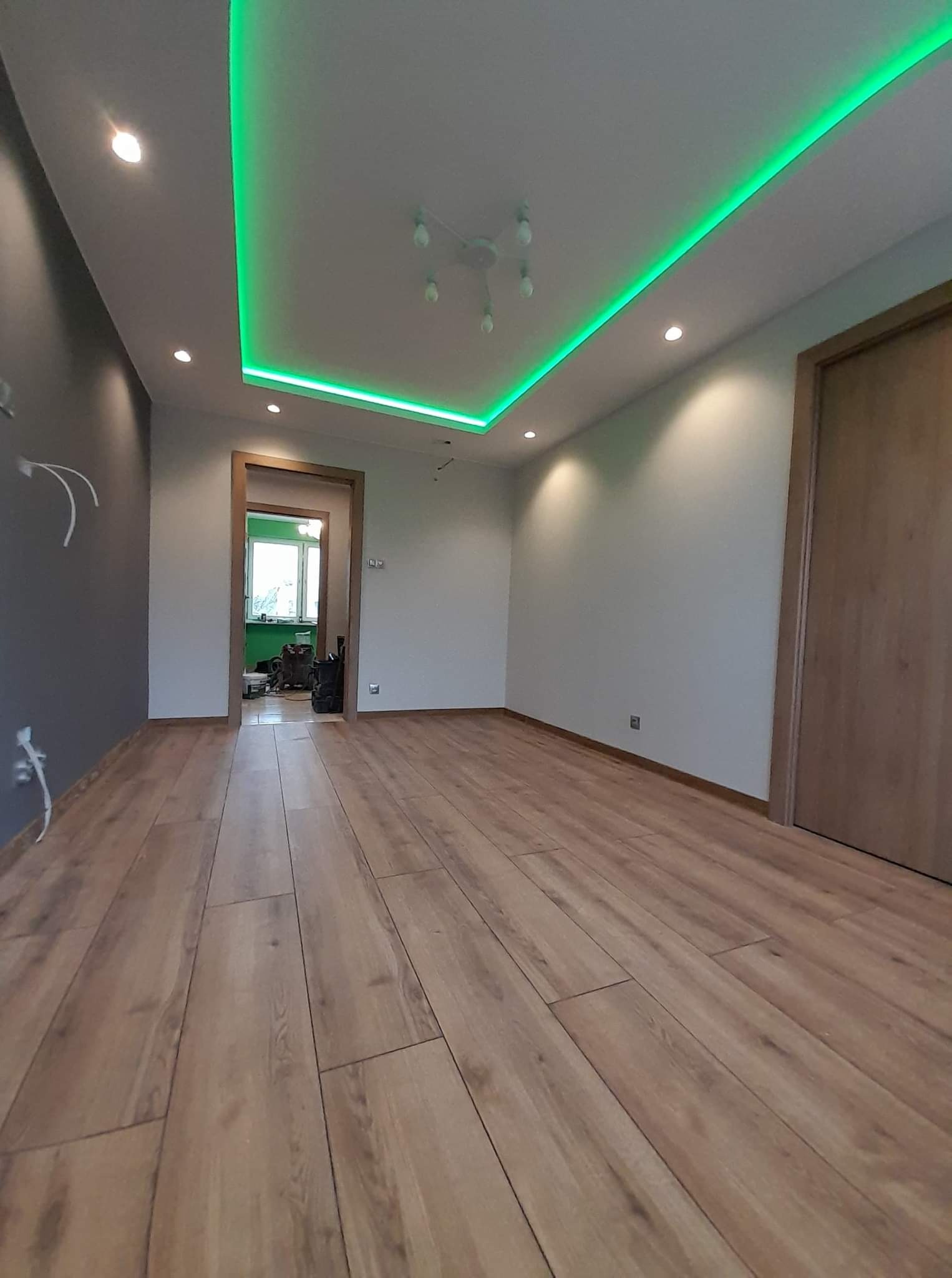 Pokój z parkietem, szarą ścianą, drewnianymi drzwiami i sufitem z oświetleniem LED w kolorze zielonym.