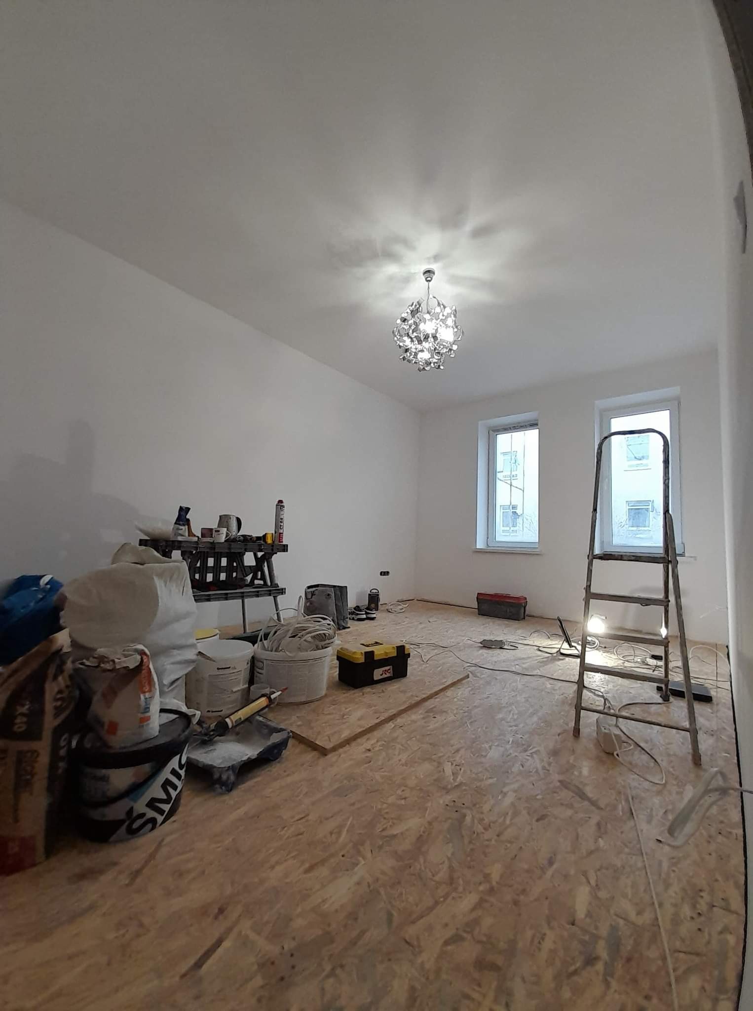 Pokój w trakcie remontu z widoczną drabiną, workami gipsu, narzędziami i zamontowanym ozdobnym żyrandolem pod sufitem.
