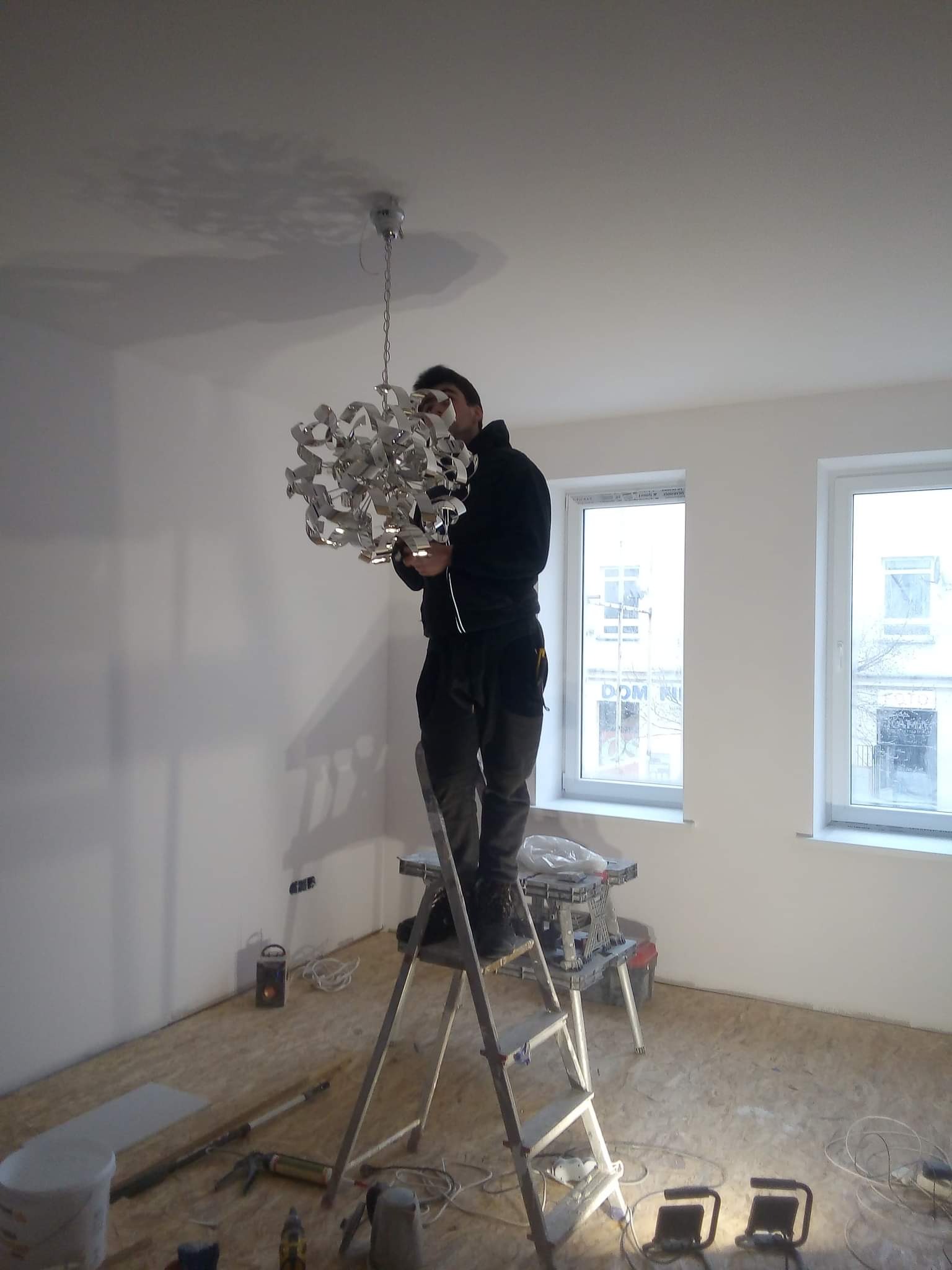 Elektryk na drabinie w nowym wnętrzu, montuje nowoczesną, srebrną lampę wiszącą z licznymi zakrzywieniami, w tle dwa okna i surowe podłoże z OSB.