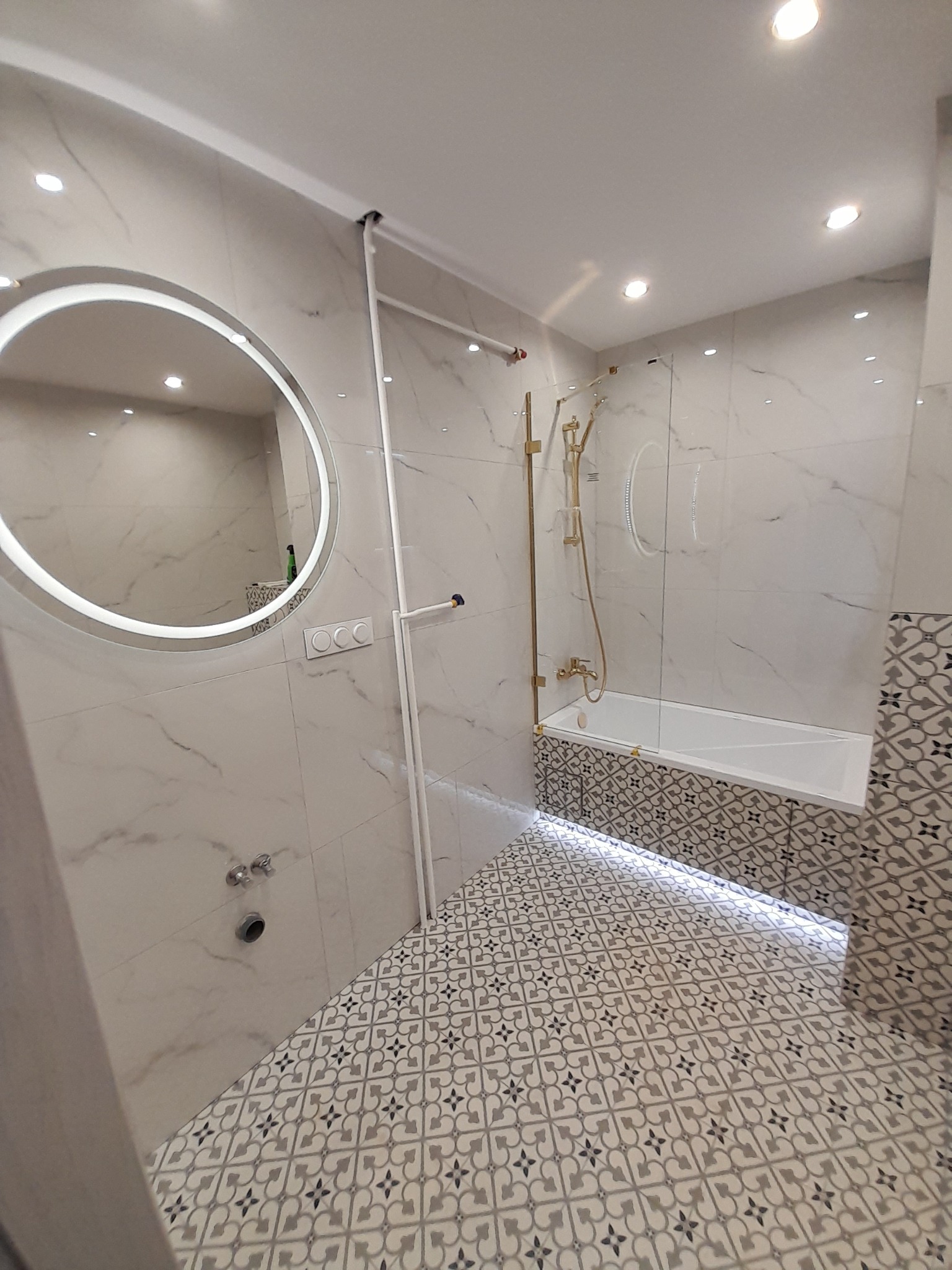 Nowoczesna łazienka z marmurowymi płytkami, okrągłym lustrem z podświetleniem LED, złotą armaturą prysznicową i podświetlaną wanną.