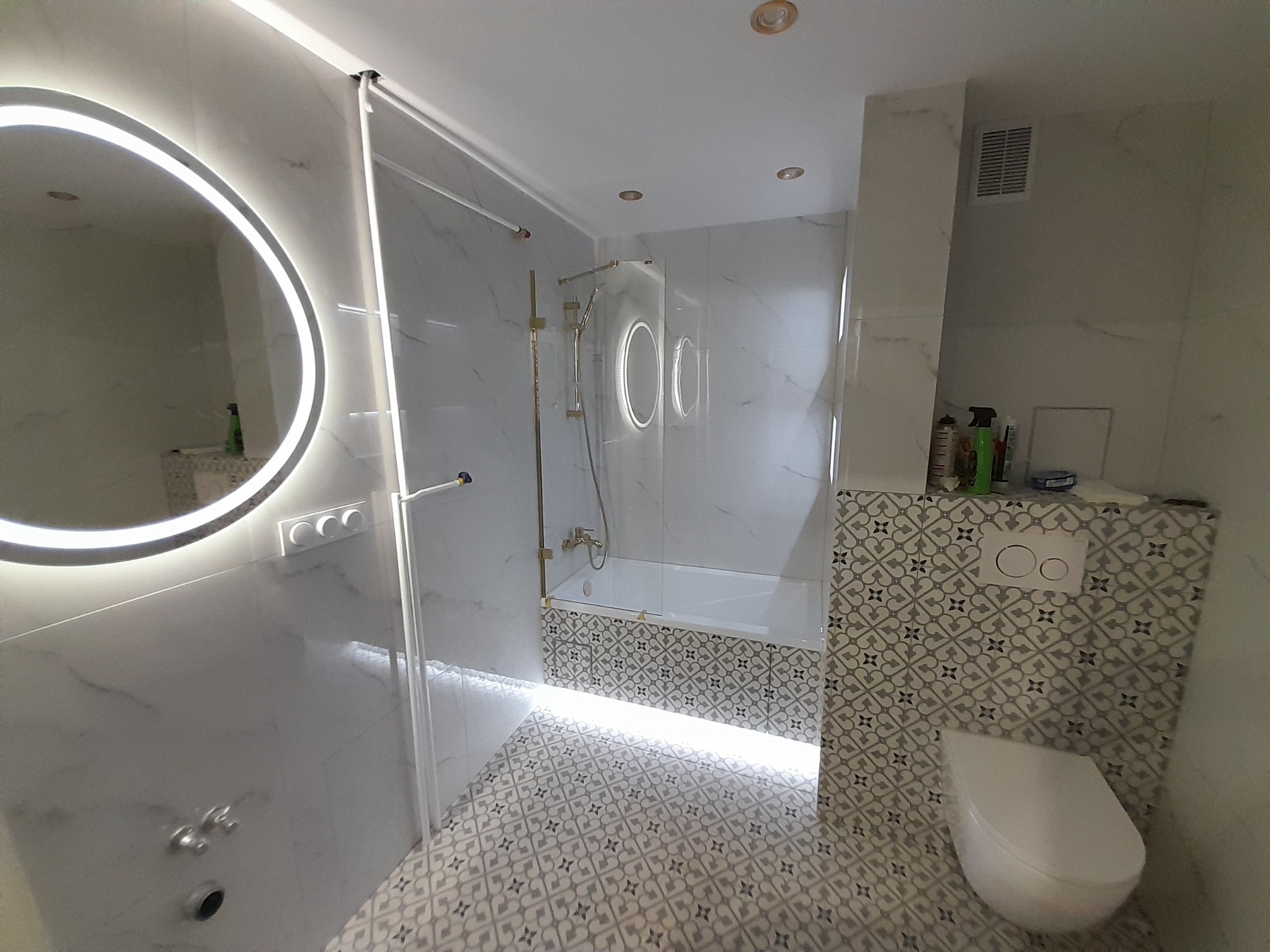 Nowoczesna łazienka z podświetlanym lustrem, kabiną prysznicową ze złotymi detalami i wzorzystymi płytkami podłogowymi.