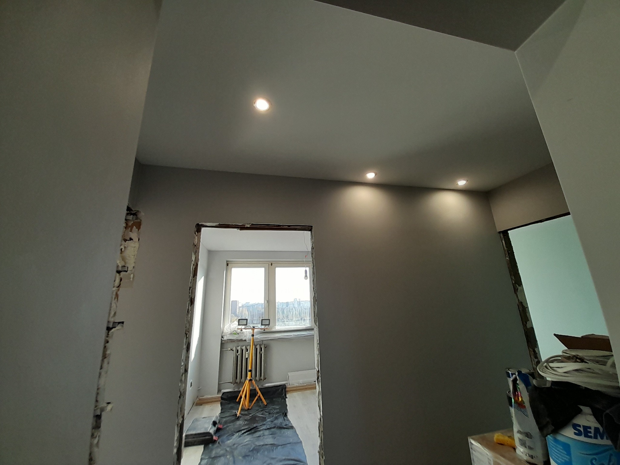 Pokój w trakcie remontu, widoczne zamontowane punktowe oświetlenie sufitowe, ściany pomalowane na szaro, widoczne przygotowania do dalszych prac wykończeniowych, lampa budowlana na trójnogu w oddali.