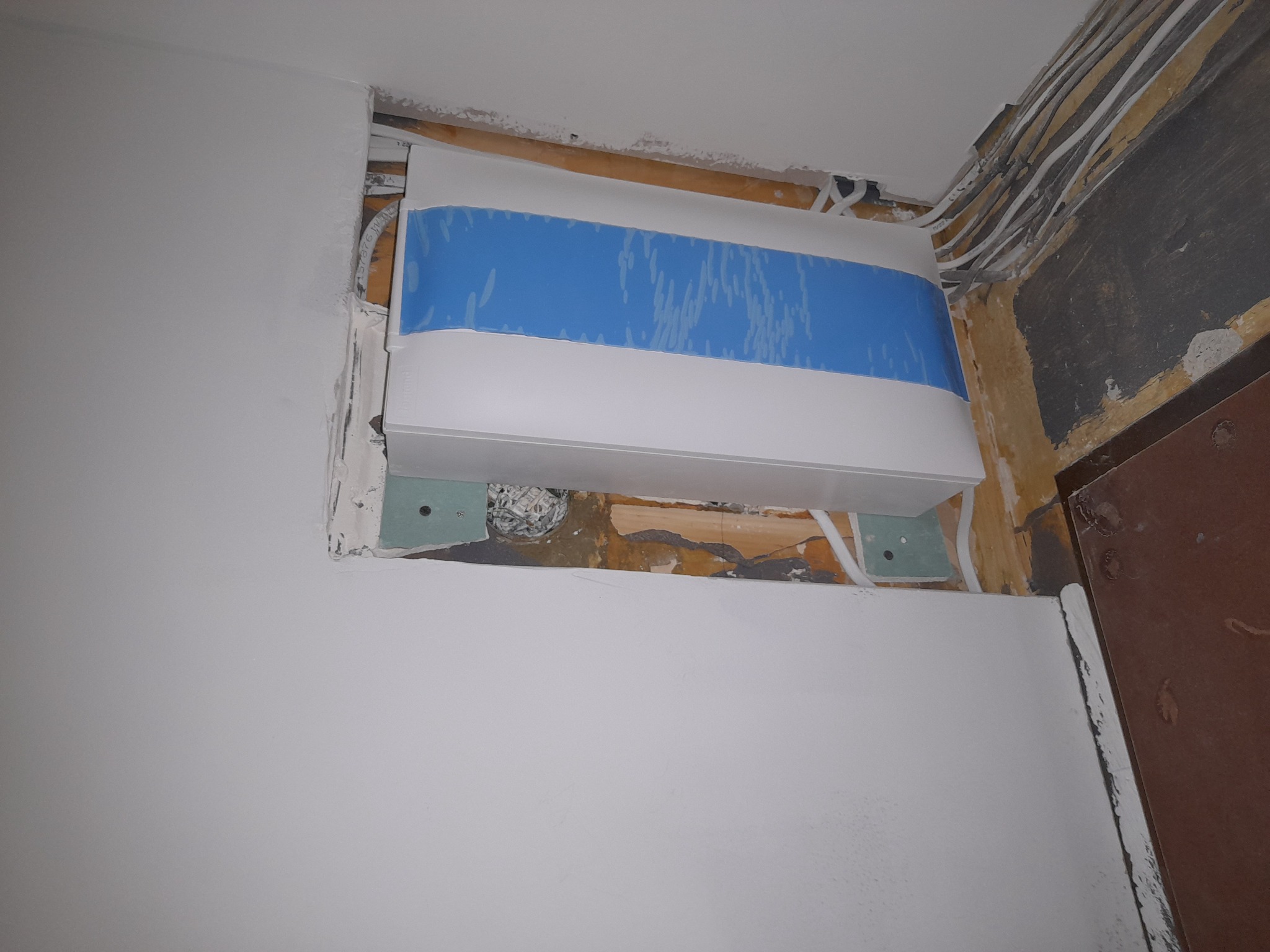 Niewykończona instalacja elektryczna: biała skrzynka z niebieską taśmą zabezpieczającą, wystające kable i przewody, ślady po remoncie na ścianach i suficie.