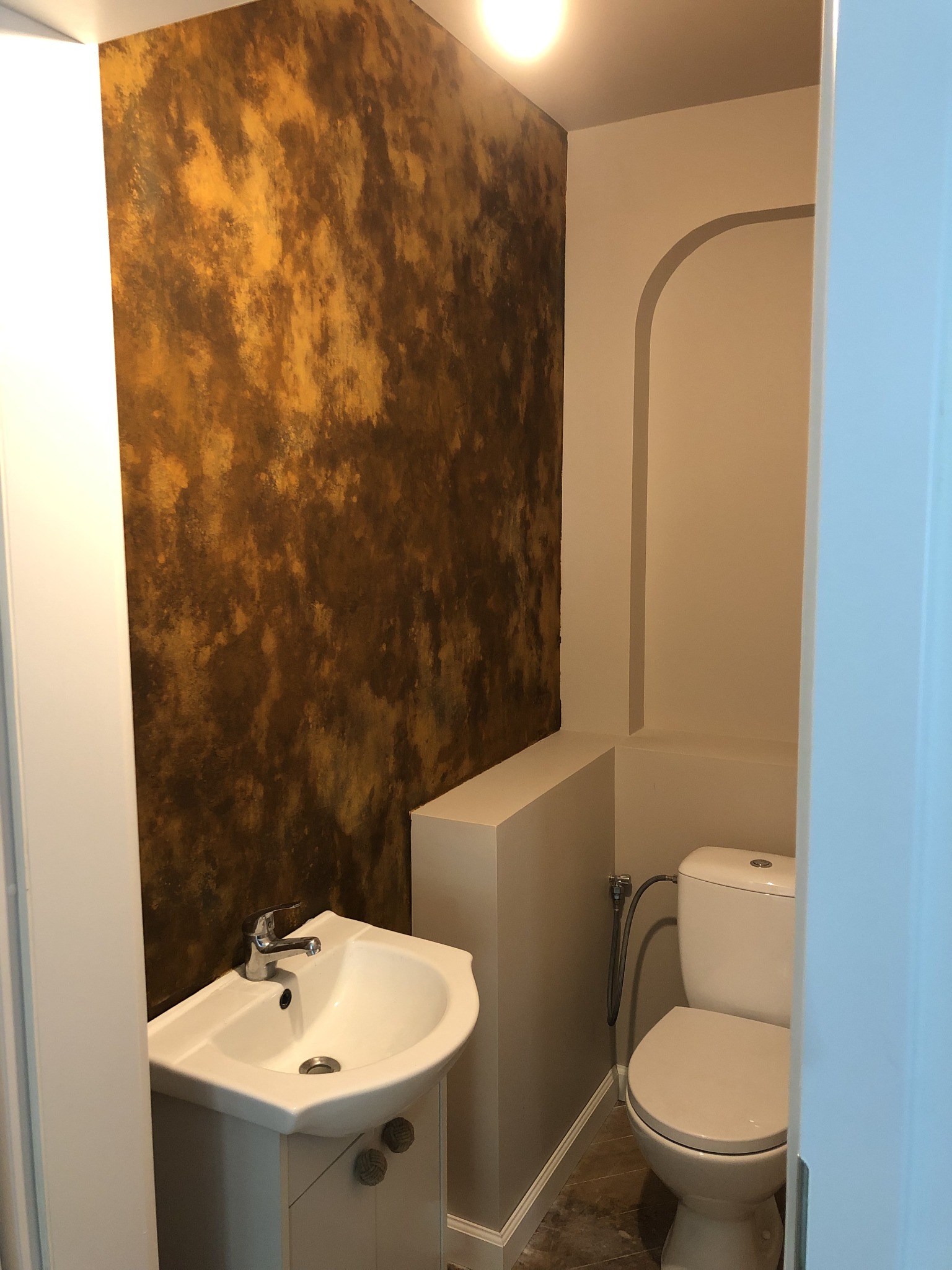 Mała łazienka z umywalką narożną i toaletą; ściana pokryta dekoracyjną farbą w odcieniach brązu i złota, imitującą marmur.