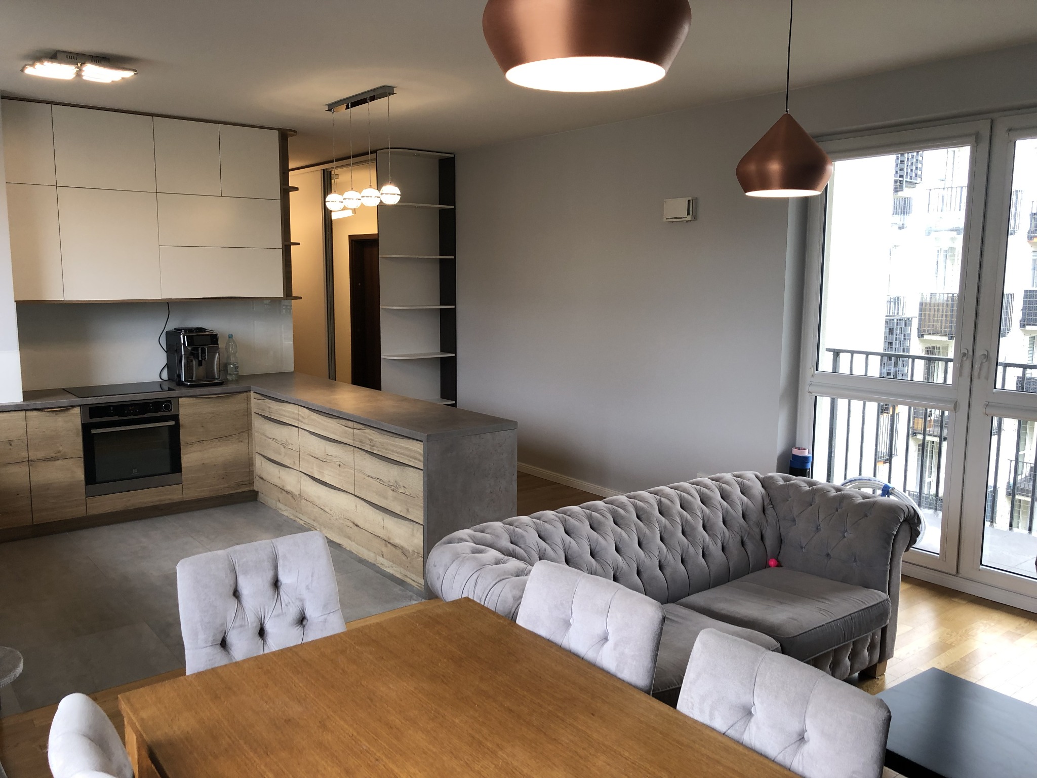 Nowoczesne wnętrze mieszkania z drewnianym stołem, szarą sofą typu chesterfield, kuchnią w odcieniach drewna i bieli oraz miedzianymi lampami wiszącymi.