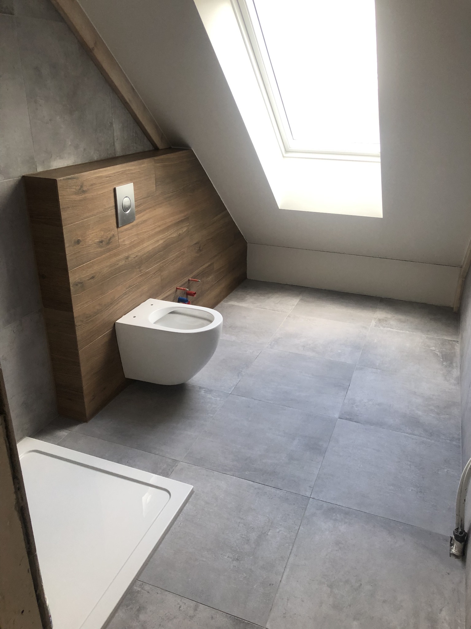 Nowoczesna łazienka na poddaszu z wiszącą toaletą na drewnianej ściance, szare płytki podłogowe i biały brodzik w rogu, doświetlona oknem dachowym.