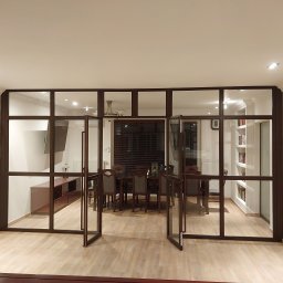 Binexus Michał Wojtaszuk - Przeszklona ściana działowa z brązowymi profilami oddzielająca dwa pomieszczenia, widok na jadalnię z zasłoniętymi żaluzjami i salon z telewizorem i regałem na książki.