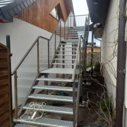 Zewnętrzne, ażurowe schody z metalu z balustradą ze szkła, prowadzące na wyższy poziom budynku, z kablem elektrycznym leżącym na stopniach.