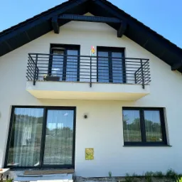 Biały dom z czarnym dachem i czarną, metalową balustradą balkonową o prostym wzorze, z kwiatami w doniczce; widoczne duże okna i drzwi balkonowe z czarnymi ramami.