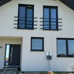 Fasada domu jednorodzinnego z czarnymi oknami zabezpieczonymi prostymi, poziomymi barierami oraz ciemnoszarymi drzwiami wejściowymi z długim, pionowym uchwytem, widoczna skrzynka na listy z numerem 5.