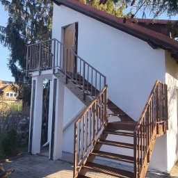 Binexus Michał Wojtaszuk - Zewnętrzne schody metalowe z balustradą prowadzące do drzwi na podwyższeniu, z widoczną konstrukcją wsporczą i otoczeniem z kostki brukowej.