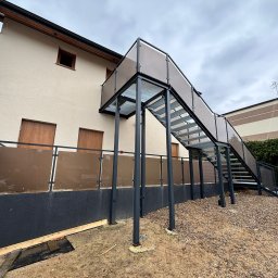 Binexus Michał Wojtaszuk - Zewnętrzne schody metalowe z balustradą ze szkła brązowego, prowadzące do drzwi na piętrze budynku. Konstrukcja stalowa w kolorze grafitowym, widok z dołu, pochmurne niebo.