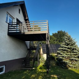 Binexus Michał Wojtaszuk - Nowoczesny, ciemnoszary metalowy balkon z poziomymi balustradami i zewnętrznymi schodami, zamontowany na jasnej elewacji domu jednorodzinnego, w słonecznym ogrodzie.