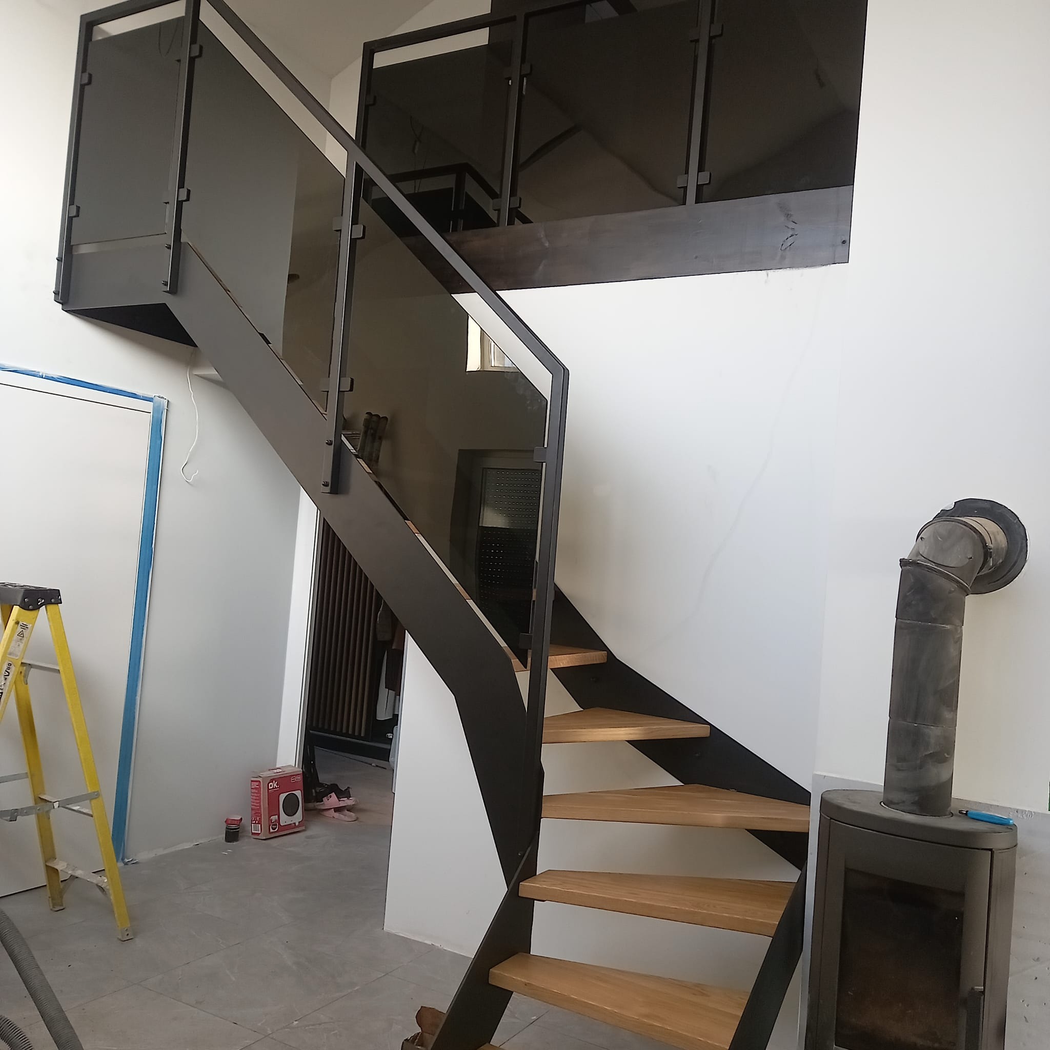 Nowoczesne schody z drewnianymi stopniami i szklanymi panelami, czarna metalowa konstrukcja i balustrady, w tle widoczny piec.