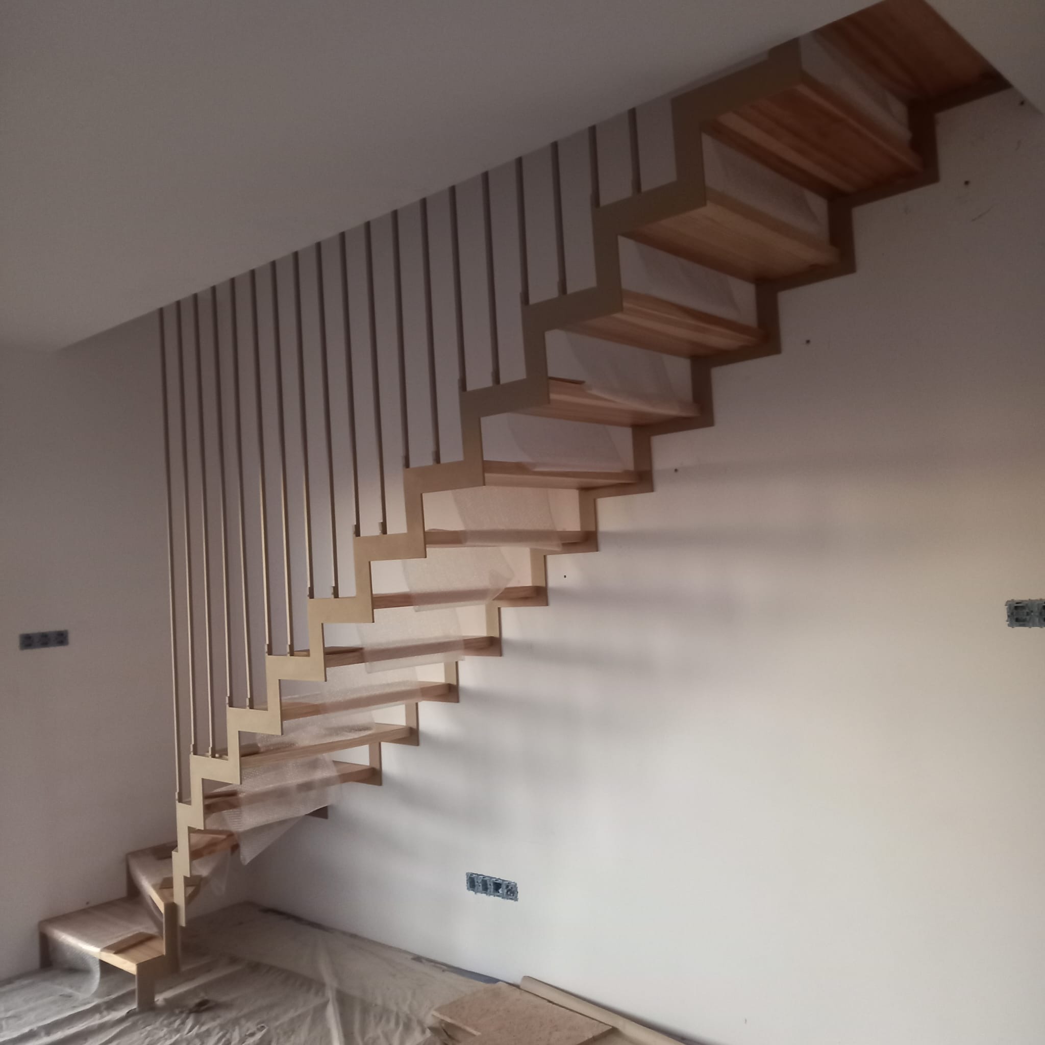 Nowoczesne schody z drewnianymi stopniami i metalową, minimalistyczną balustradą z pionowych prętów, częściowo zabezpieczone folią ochronną, w trakcie montażu w surowym wnętrzu.