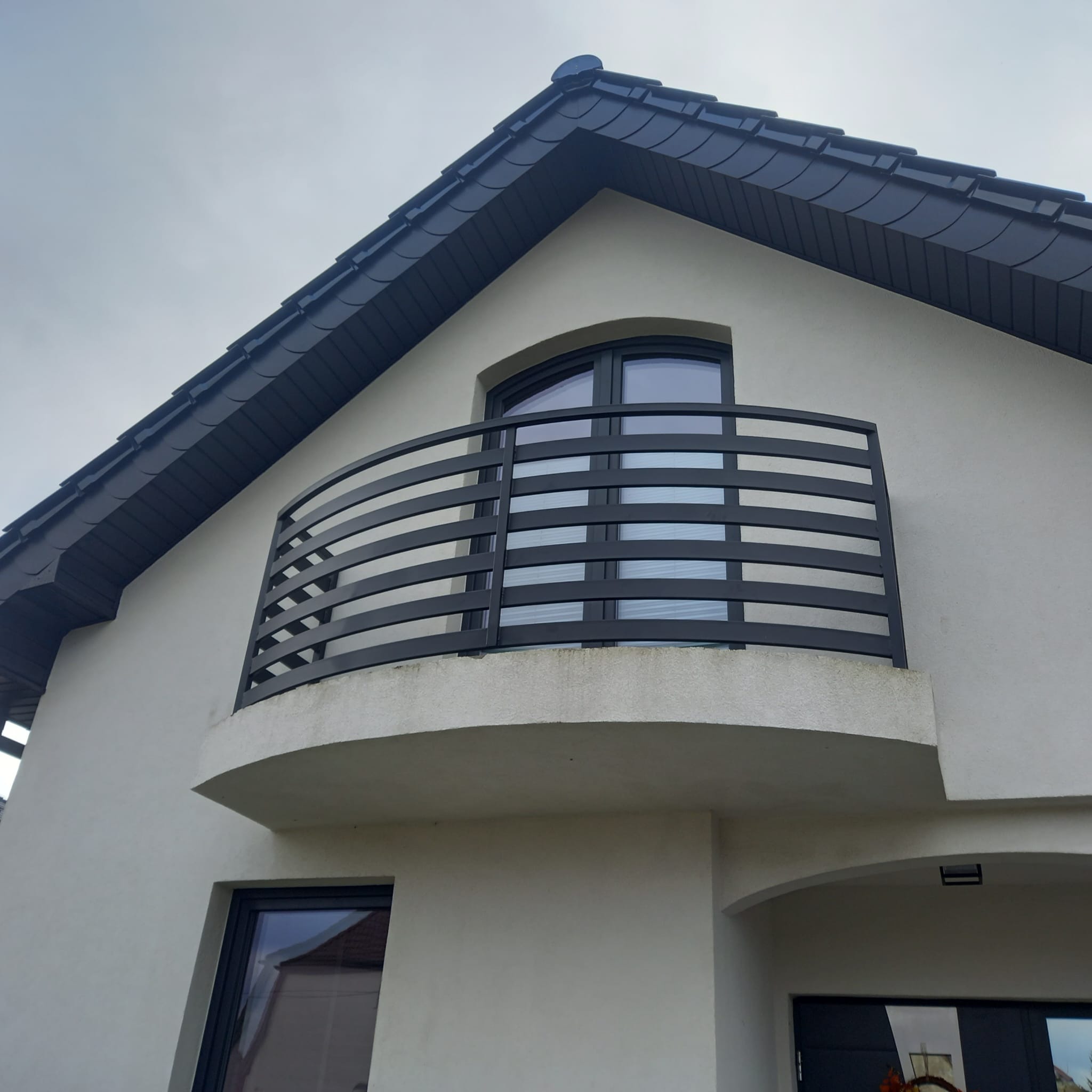 Nowoczesny balkon z czarną, poziomą balustradą na tle jasnej elewacji domu z ciemnym dachem, widok z dołu.