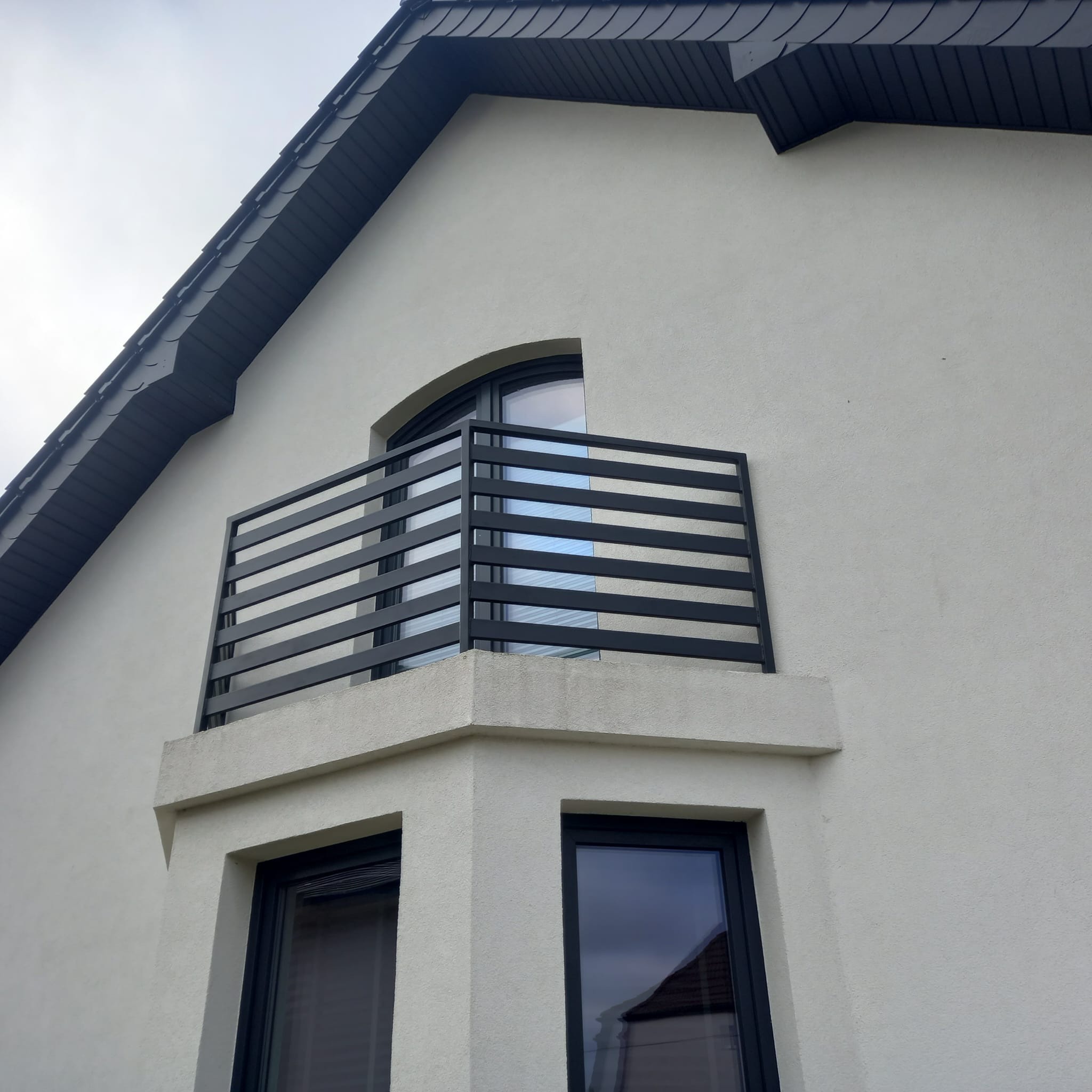 Czarna, pozioma balustrada metalowa na balkonie z oknem łukowym, widok z dołu na elewację budynku z szarym dachem.